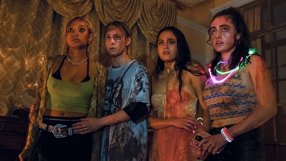 A shot from the movie Bodies Bodies Bodies shows four frightened looking, bloodied young women. One of them is wearing a bunch of glowsticks.