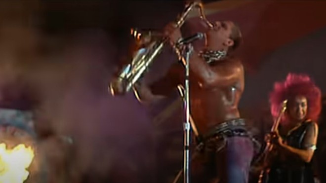 A screenshot from the movie Lost Boys shows a muscular, shirtless man, oiled skin glistening, thick chain wrapped around his neck, passionately playing a saxophone