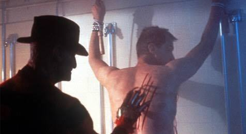 A screenshot from Nightmare on Elm Street Part 2 shows Freddy Kruger using his clawed glove to cut the back of a shirtless man who is tied up in a shower